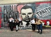 Four people stand in front of a colorful mural featuring a large portrait and various text, including "Public Enemy" and "John Dillinger.