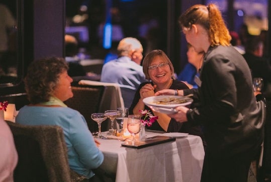 Tow women smiling happily while food is being served by a waitress wearing all black on the Chicago Premier Dinner Cruise on Lake Michigan.