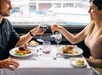 A couple cheering their glasses together over their dinners with a large window behind them letting light in on the Chicago Premier Dinner Cruise on Lake Michigan.
