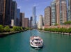 A white tour boat with passengers travels along a river lined with modern high-rise buildings on a clear day in an urban cityscape.