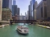 A tour boat travels along the Chicago River, surrounded by tall skyscrapers and a bridge, with people visible on the boat's deck.