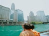 A person wearing sunglasses and an orange jacket looks at tall buildings along a river in a city.
