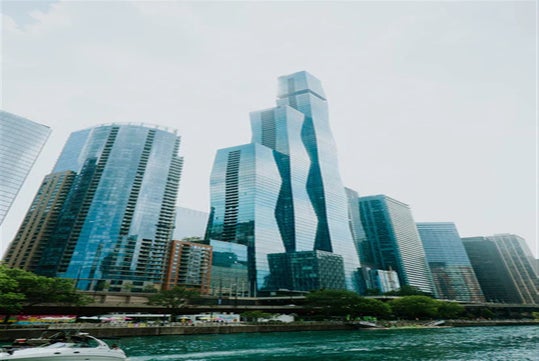 Tall, modern glass skyscrapers line a city waterfront on a cloudy day, with one uniquely patterned tower standing out among surrounding buildings. A boat is visible in the water.