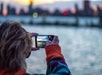 A person wearing a colorful jacket takes a photo of a city skyline at sunset across a body of water using a smartphone.