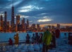 People stand on a boat deck at dusk, photographing and observing the illuminated city skyline across the water.