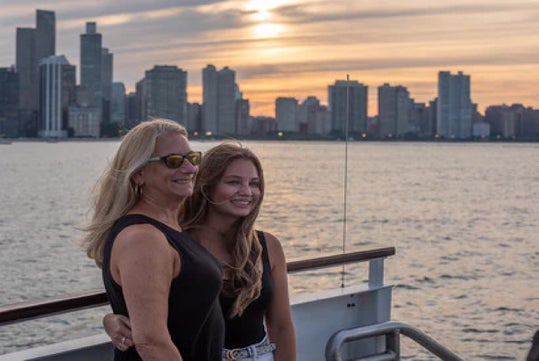 Two women stand side by side on a boat at sunset, with city skyscrapers and water in the background.