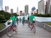 Cyclists in green shirts ride red bikes on a wooden bridge in Millennium Park, with Chicago’s skyline in the background.