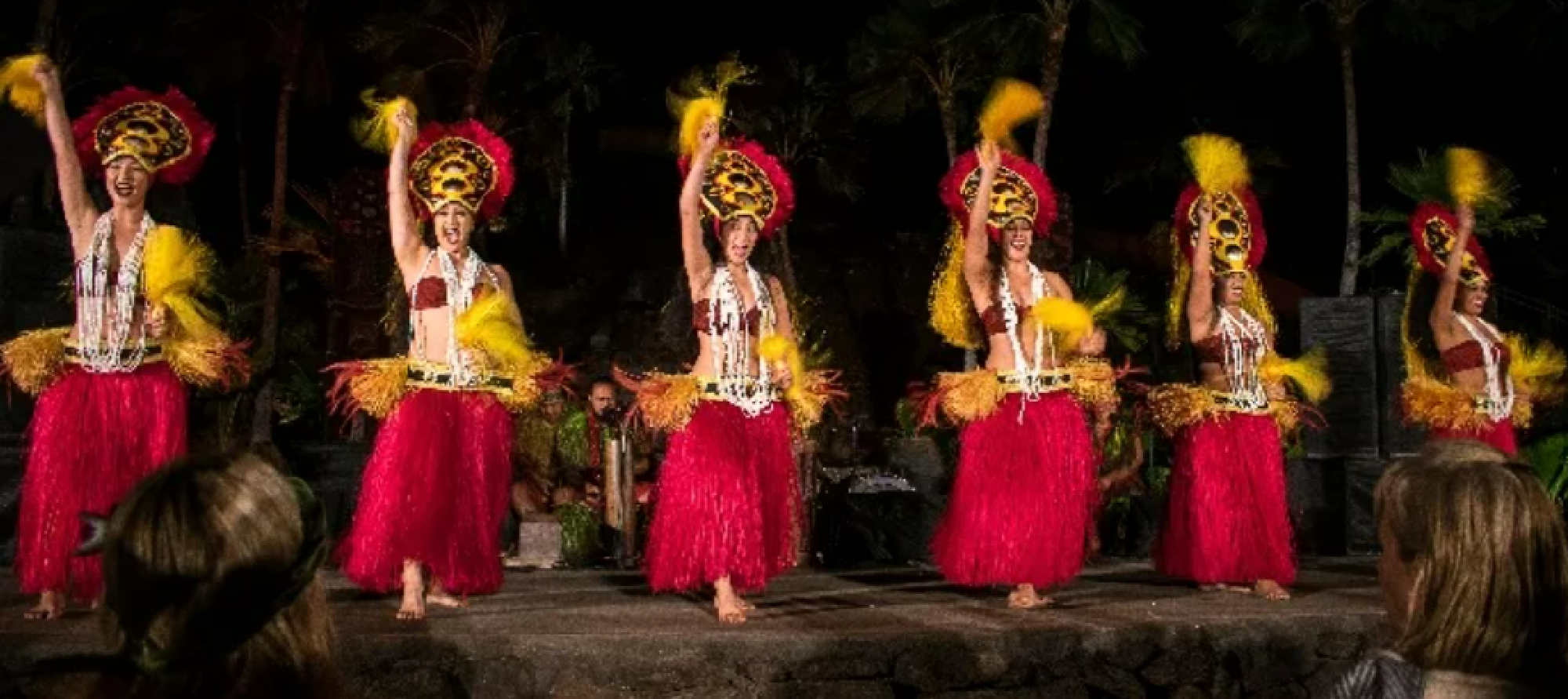 Five dancers perform in traditional grass skirts and headdresses at a nighttime outdoor event, with audience members visible in the foreground.