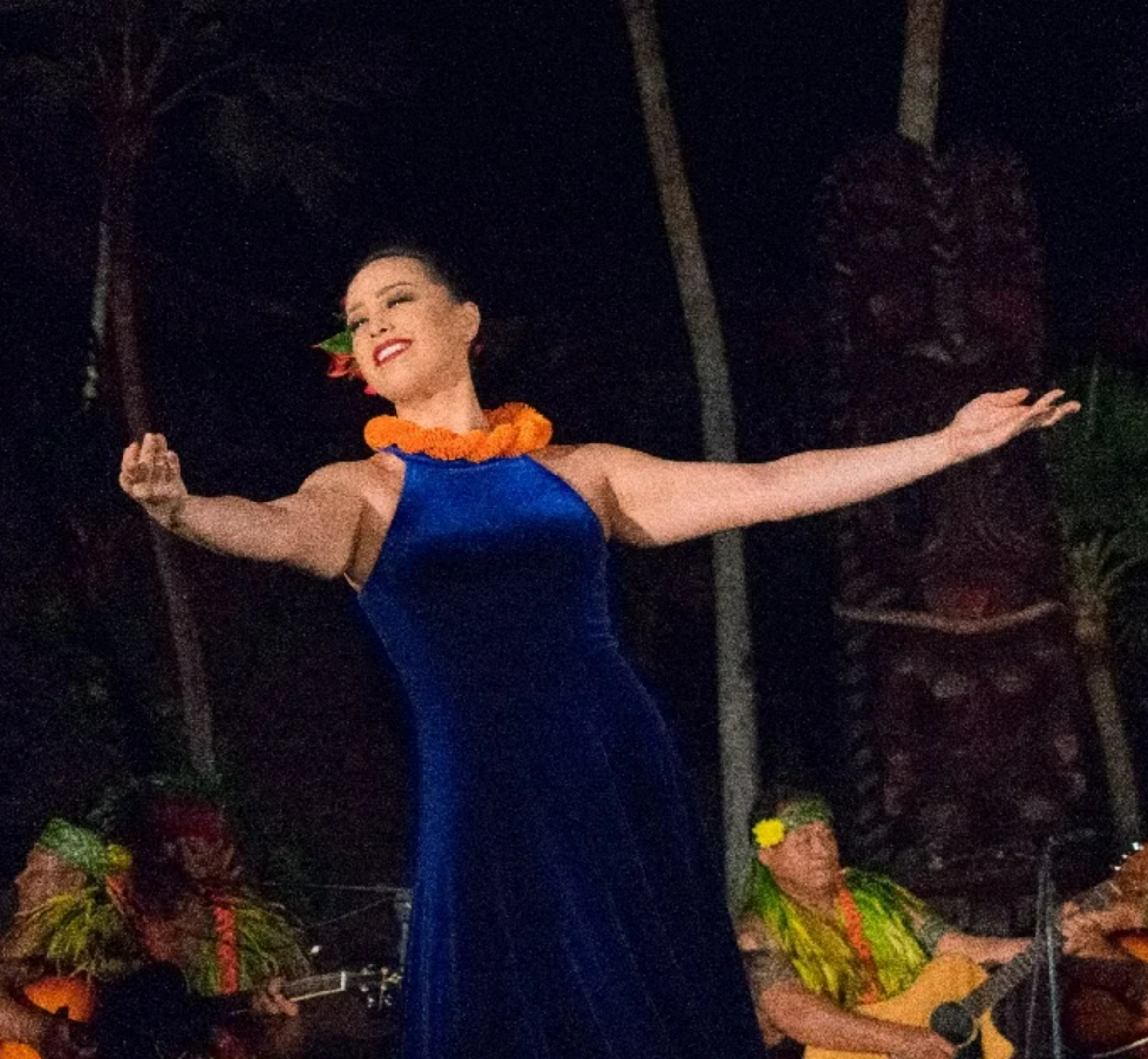A woman in a blue dress performs a dance with her arms outstretched, wearing an orange lei. Musicians in traditional attire play string instruments in the background.