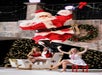 Santa Claus is mid-jump in front of a sleigh with three women in festive outfits, with Christmas trees and holiday decorations in the background.