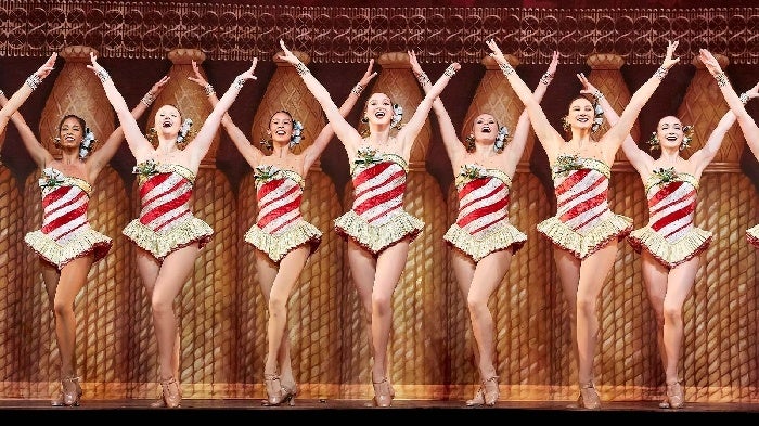 Close up of a few of the Radio City Rockettes in red and white stripped costumes with both of their arms up on stage.