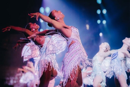 Close up of a Rockette wearing a silver costume with small wings on the back with other matching dancer in the background.