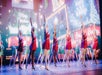 The Radio City Rockettes performing in red sparkling dresses with one of their arms up and Christmas cards on the screens behind them.
