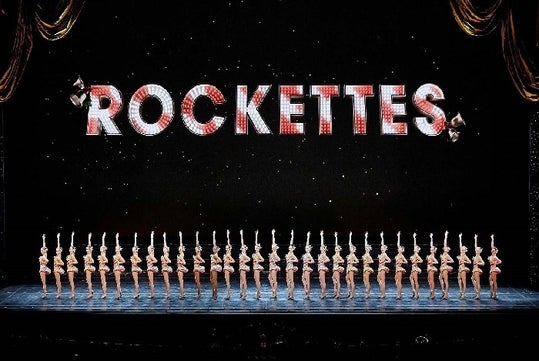 The Radio City Rockettes in silver and red costumes on stage in a line with one of their arms up and a sign saying "ROCKETTES" over them.