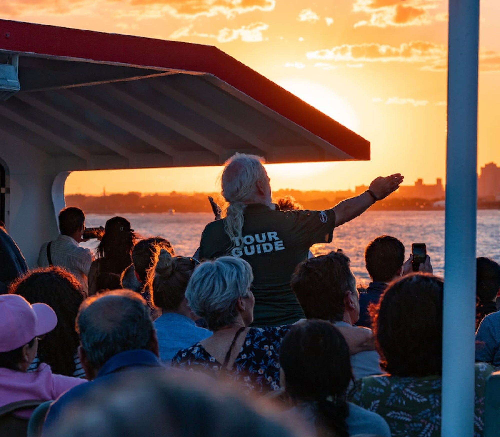 A tour guide gestures toward the sunset as a crowd of people observe from the deck of a boat.