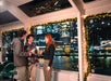 Three passengers relax inside the boat's spacious main cabin, with refreshing drinks in hand, as they chat and enjoy the breathtaking views of the Hudson River and New York skyline through the large windows in the background