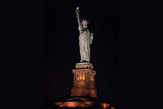 The Statue of Liberty is illuminated at night, standing on its pedestal against a dark sky.