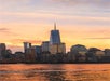 Skyline of a city with a tall modern skyscraper at sunset, reflecting warm orange hues on the calm water in the foreground.