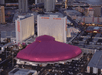 Aerial view of the Las Vegas hotel and casino complex with a large, pink, heart-shaped roof and surrounding high-rise buildings and parking lots.