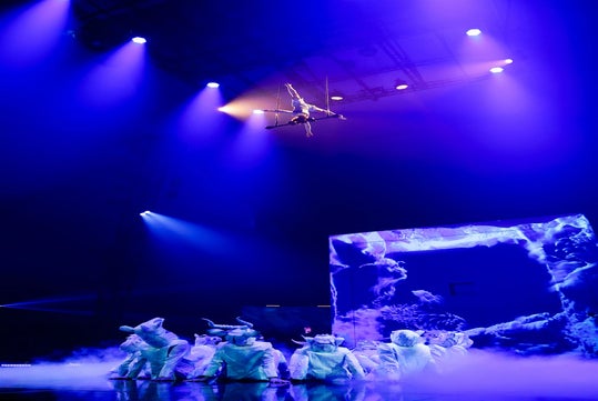 A performer suspended in the air above a group of dancers in animal costumes on a foggy stage, lit by purple-blue stage lights.