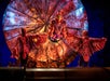 Three performers in red bird costumes pose on stage in front of a large, circular, fan-like backdrop with dramatic lighting.