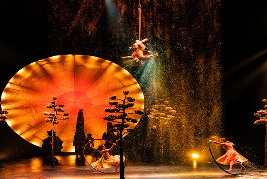 Aerial performer hangs upside down above a stage with a glowing circular backdrop, water falling, and two dancers posing in illuminated hoops below.