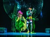 Performers in colorful, whimsical costumes resembling plants and sea creatures stand on stage with a starry backdrop, engaging in a lively theatrical performance.