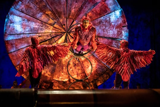 Three performers in red bird costumes pose on stage in front of a large, circular, copper-toned backdrop; one performer perches on a metal ring structure.
