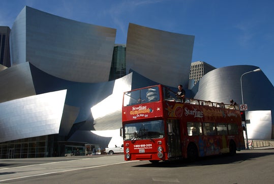 A red double-decker tour bus drives past the silver, angular exterior of the Walt Disney Concert Hall in Los Angeles on a sunny day.