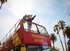 Two people on the upper deck of a red CitySightseeing Los Angeles tour bus look at a map and point, with palm trees and a bright sky in the background.