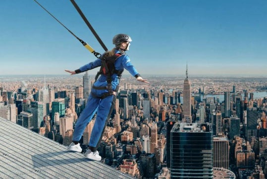 Person in a safety harness leans forward from a high platform overlooking a city skyline.