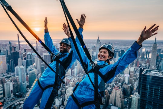 Two people in blue jumpsuits and safety harnesses pose with arms raised on a high-altitude outdoor sky deck above a city skyline at sunset.
