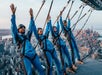 Four people in blue suits and helmets suspended on a platform high above a city skyline, with arms outstretched.