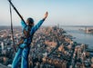 Person wearing a safety harness stands on a high platform with arms raised, overlooking New York City and the surrounding skyline at sunset.