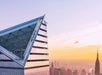 Close-up of a modern skyscraper's angled glass facade at sunset, with cityscape and another tall building in the background.
