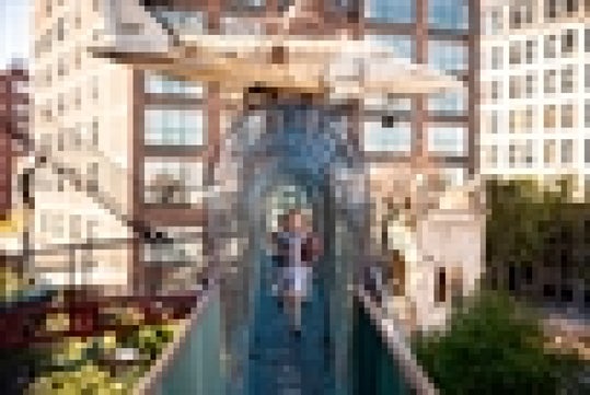A child walks through a transparent tunnel bridge connecting two buildings in an urban outdoor setting.