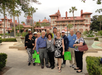 A group of adults poses together outdoors in front of a historic building with red roofs, lush greenery, and a fountain nearby.