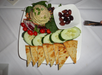 A plate with pita bread slices, cucumber, cherry tomatoes, leafy greens, a scoop of hummus, and a small dish of assorted olives on a white tablecloth.
