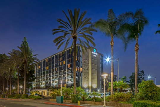 A multi-story hotel building with illuminated signs, surrounded by tall palm trees, against a twilight sky.