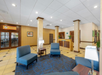 Hotel lobby with blue chairs, small tables, beige walls, and a reception desk. Ceiling lights illuminate the area, and a doorway is visible in the back.