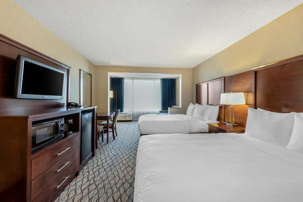 Hotel room with two double beds, a wall-mounted TV, a desk with a chair, and a window with dark curtains.