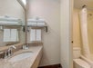 Bathroom with a sink, mirror, and toiletries on the counter. Towels are stacked on a shelf above two hanging towels. A shower with a curtain is next to the toilet.