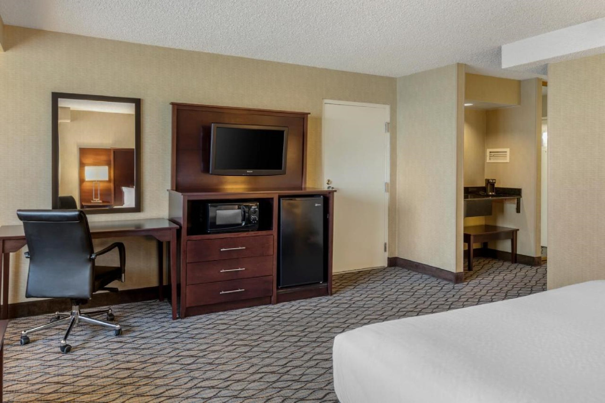 Hotel room with a desk, chair, TV, microwave, and mini-fridge. A bathroom is partially visible in the background.