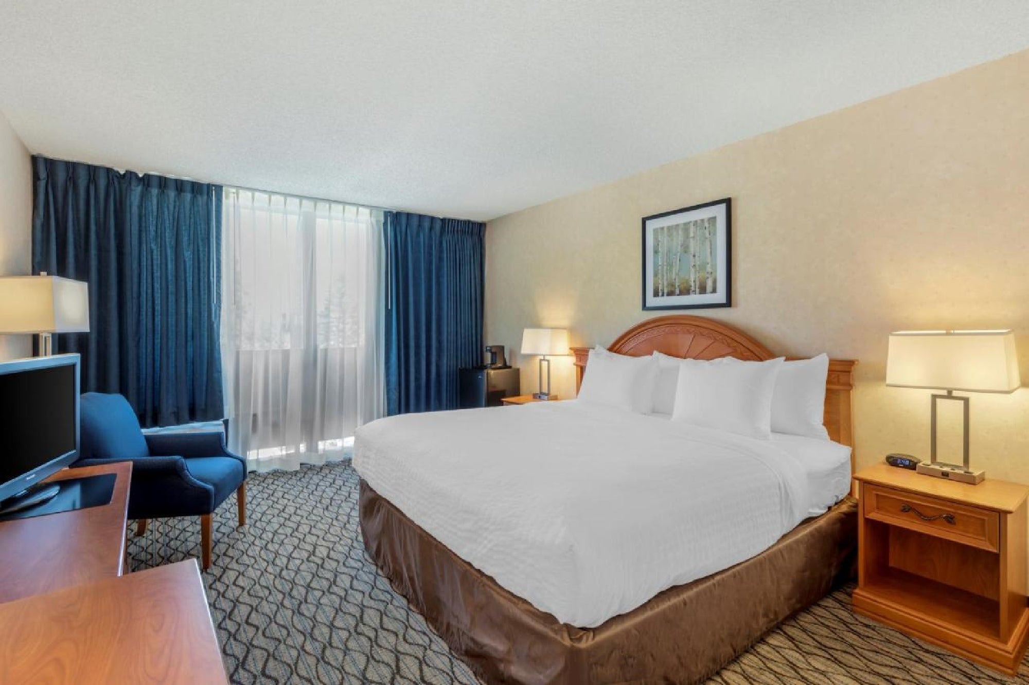 Hotel room with a large bed, two lamps, an armchair, a desk, and a window with blue curtains. Walls are beige and carpet is patterned.