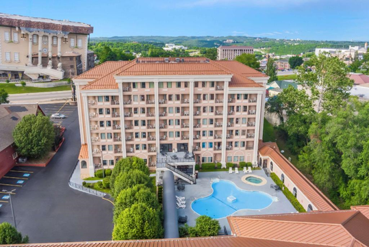 A multi-story hotel with a terracotta roof, outdoor pool, and surrounding greenery, viewed from above on a clear day.