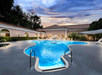 Outdoor swimming pool at a hotel or resort, surrounded by lounge chairs and trees, with a building and sunset sky in the background.