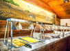 Buffet counter with trays of cut fruit and plates, set against a wooden wall with a mural of zebras and African scenery.