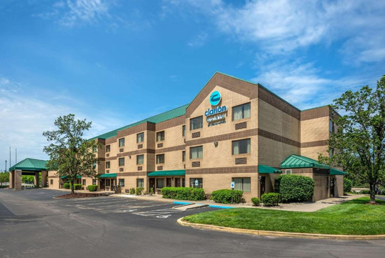 A three-story hotel building with a green roof and parking area in front, under a partly cloudy blue sky.