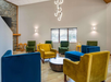 A modern lounge with colorful velvet armchairs, including blue, yellow, and green, arranged around a round wooden table. A contemporary chandelier hangs overhead.