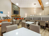A modern dining area with tables and chairs, featuring a stone fireplace, a mounted TV, and a coffee station on the left. Contemporary lighting hangs from the ceiling.